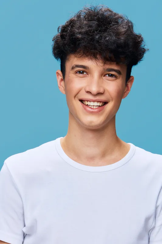 smiling young man with curly hair wearing white shirt on blue background, natural light, casual style, happy emotion, studio portrait