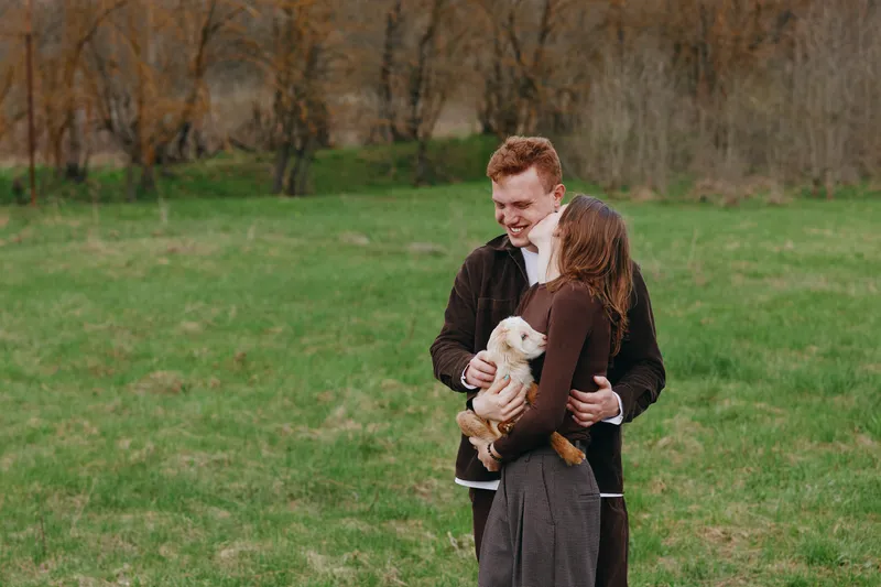 Young couple embraces outdoors while holding a small white baby goat in a green field during autumn, sharing a joyful and tender moment together.