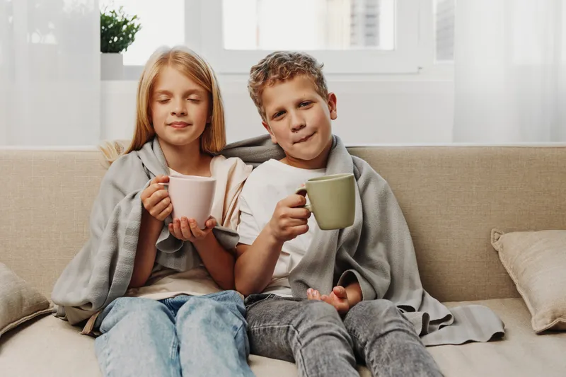 Two children wrapped in cozy blankets enjoy warm drinks while sitting on a sofa in a bright living room, expressing comfort and relaxation during a calm moment at home.