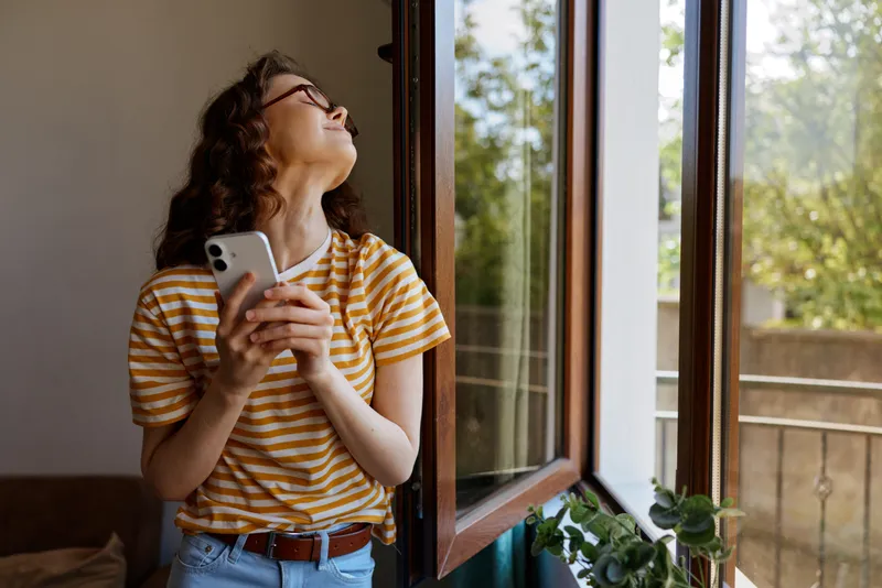 A woman in a striped t shirt stands by an open window, looking up with a thoughtful smile, phone in hand. Bright home scene, casual mood, reflective moment in the morning light softly.