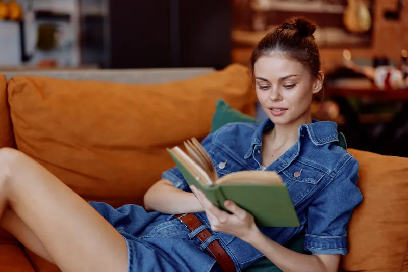 Молодая женщина наслаждается тихим моментом, читая книгу, одетая в джинсовое платье, сидя на оранжевом диване в уютной гостиной