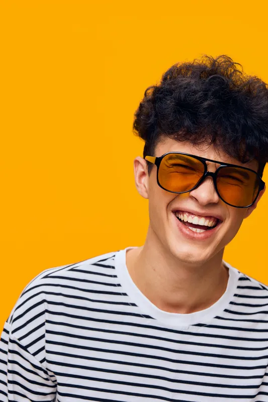 smiling man wearing striped shirt and orange sunglasses laughing cheerfully on vibrant yellow background, youthful and energetic portrait with casual style