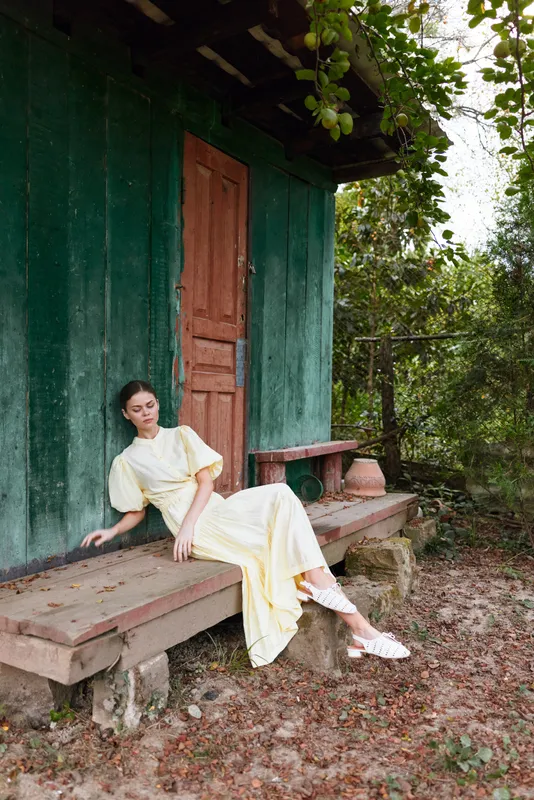 A woman in a pale yellow dress sits calmly on a wooden step beside a green rustic house doorway, enjoying outdoor garden surroundings and soft daylight.