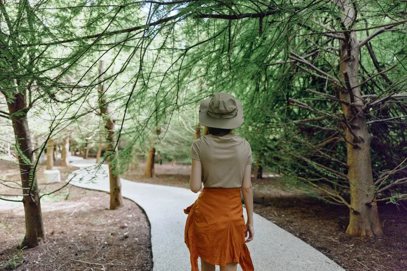 Woman walking on a winding forest path under tall trees, wearing a sun hat and orange skirt, back view captures peaceful trail, nature stroll and outdoor exploration in daylight.