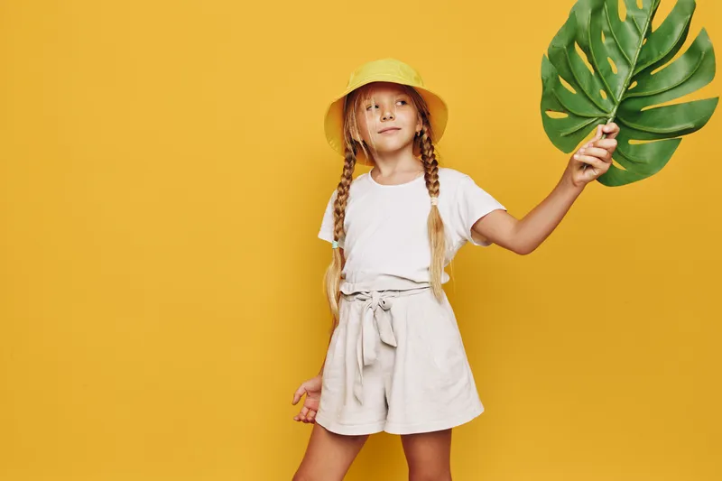 Young girl with braids wearing a yellow hat and white outfit posing confidently against bright yellow background. Studio portrait with strong contrast and summer theme.