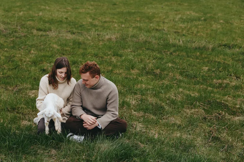 Lifestyle scene with a couple outdoors, a goat kid nearby on the grass, wearing cozy sweaters, relaxed mood, natural light, candid moment of gentle affection in a calm countryside setting