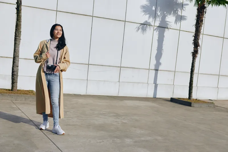 Fashion, streetstyle, coat, outdoors, sunlight, woman, casual, urban, scene with beige trench and denim, bag slung, confident stance amid modern architecture