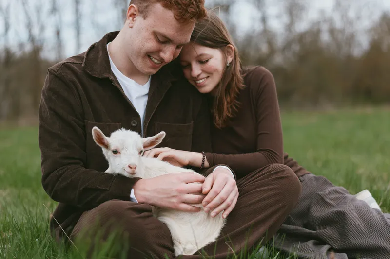 A couple sits on the grass in a sunny pasture, gently stroking a white goat kid, as curious animals peek over the fence. A countryside lifestyle moment full of affection and companionship.