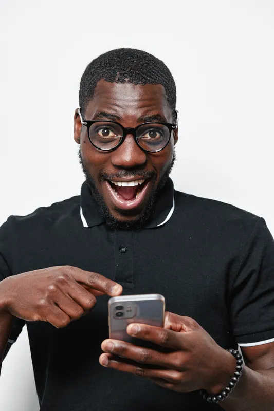 Black man with dark skin and glasses wearing black polo shirt smiling excitedly while using smartphone isolated on white background. African ethnicity, technology and communication concept.