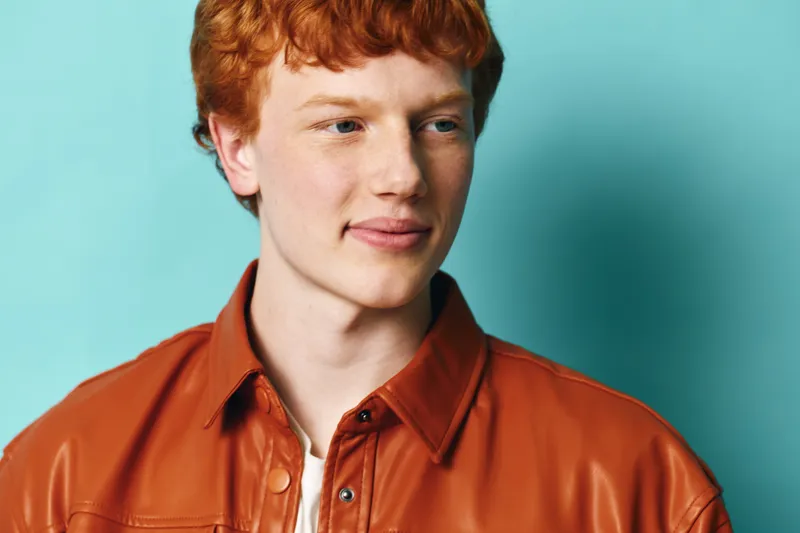 Young man with red hair in brown leather jacket looking sideways on blue background. Portrait, casual style, bright natural light with thoughtful expression and clear skin tone.