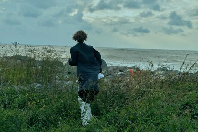 Runner coastline sea shore outdoors landscape movement weather, a windswept scene along the coast with grasses in the foreground and an overcast sky, highlighting outdoor motion and nature immersion