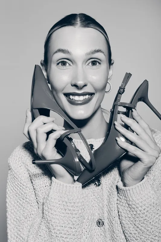 Smiling caucasian woman with light skin holds high heel shoes near face. Concept for fashion, footwear, style, happiness, and shopping enjoyment.