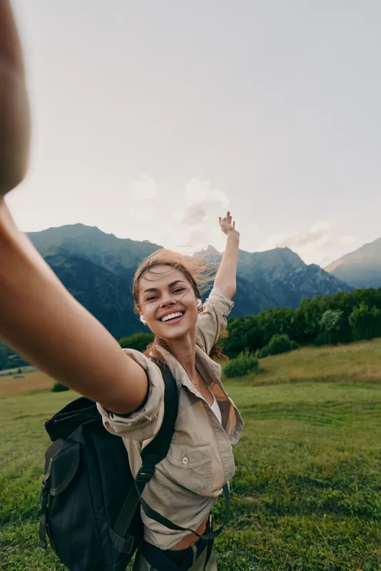 Joyful woman with backpack outdoors, reaching up in a sunlit landscape with mountains in the background, reflecting a carefree lifestyle and travel vibe.