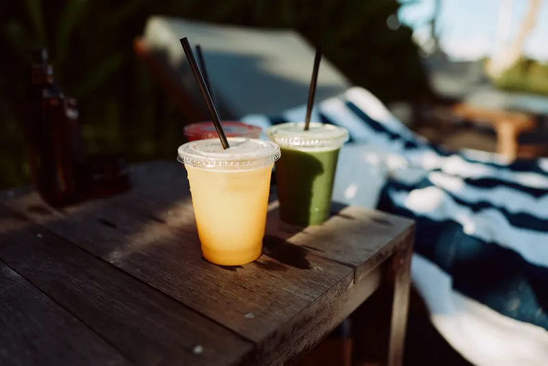 Two cold drinks on a rustic wooden table outdoors, orange smoothie and green juice, tall cups with straws, bright sun overhead, blurred campsite backdrop, calm relaxing scene
