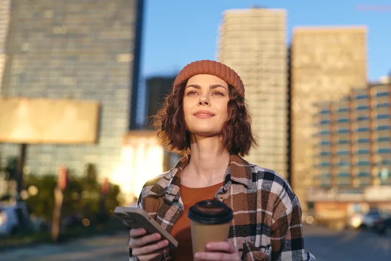 Woman holding coffee and phone in city street, smile warmed by sunlight during golden hour glow, candid lifestyle portrait capturing authenticity, mindful living and emotional storytelling.