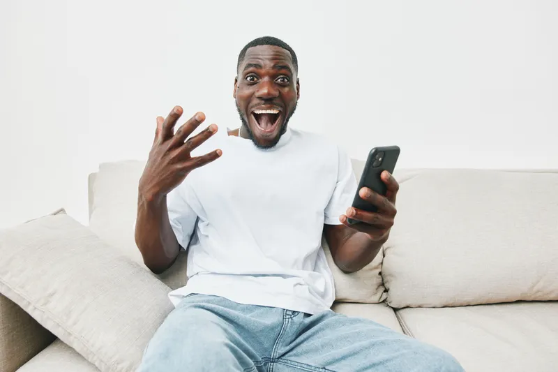 Happy young African man sitting on a couch, excitedly using a smartphone in a modern, minimalist living room with light colors, reflecting joy and connectivity