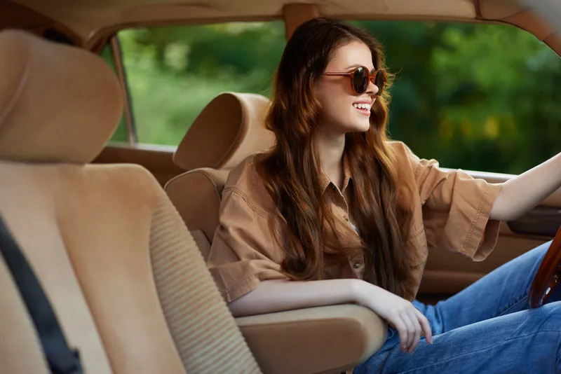 Young woman smiling in a beige car interior, wearing casual clothing and sunglasses, enjoying a bright, sunny day, reflecting confidence and freedom.