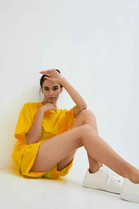 Woman sitting wearing bright yellow dress and white sneakers with relaxed pose against a white background, projecting casual fashion and modern minimalism style.