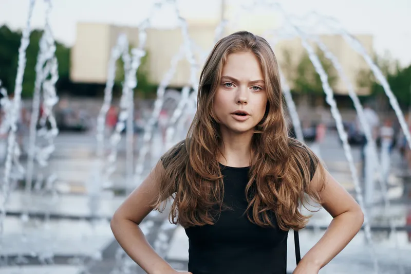 Женщина у фонтана на улице в чёрной футболке, обрезанный вид, распущенные волосы, красивая модель
