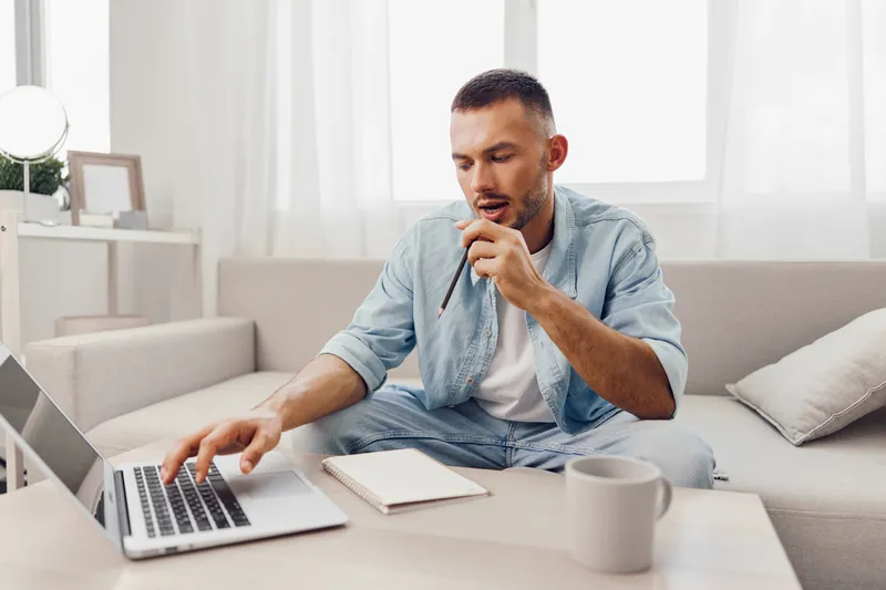 Focused man engaged in remote work, sitting at a modern desk with a laptop, notebook, and coffee cup, showcasing productivity and contemporary lifestyle trends.
