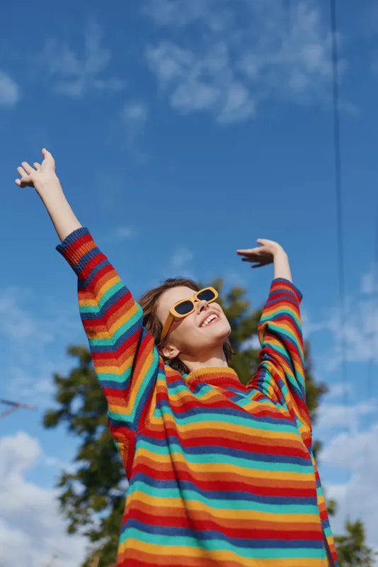 Young woman in a colorful striped sweater with arms raised, smiling and wearing yellow sunglasses under a clear blue sky. Early 20s casual outfit, joyful outdoor lifestyle portrait.