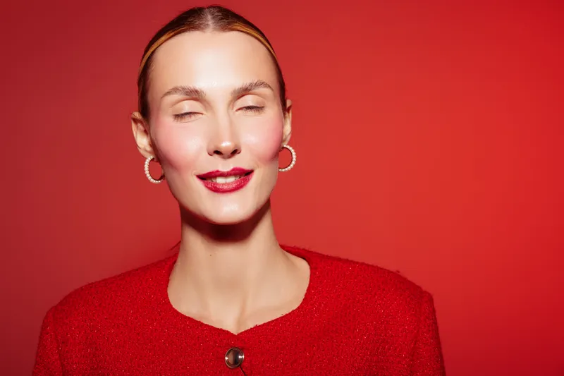 Close-up of a smiling woman with closed eyes wearing red lipstick and red sweater against red background. Concept of beauty, fashion, and confident female portrait.