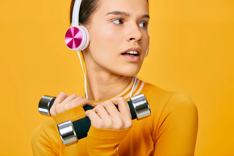 Woman in vibrant yellow athletic wear lifts silver dumbbells while wearing pink headphones, mid workout with a focused expression, bright studio backdrop, embodying a dynamic fitness moment.