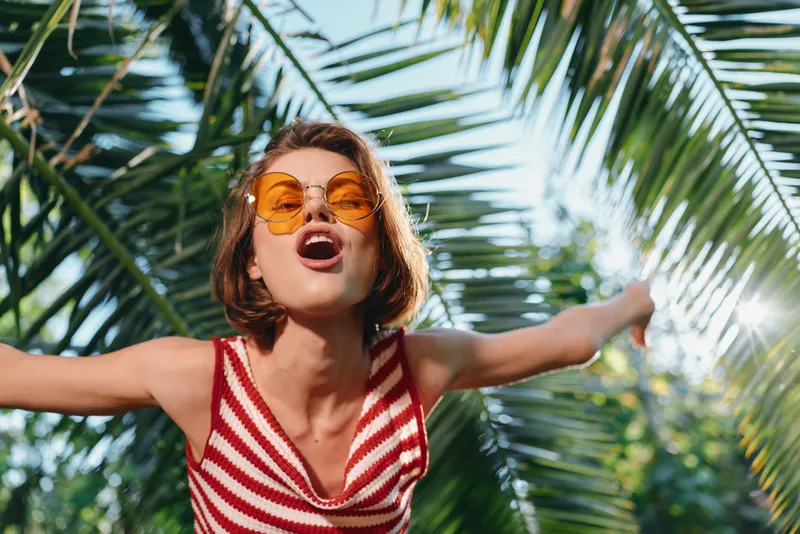 A cheerful young woman wears a striped red and white top with orange sunglasses, arms wide, enjoying a sunny tropical day among lush palm trees, exuding freedom, energy, and playful confidence.