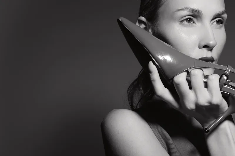 Young white woman with pale skin holds high heel shoe near face in black and white minimalist portrait.