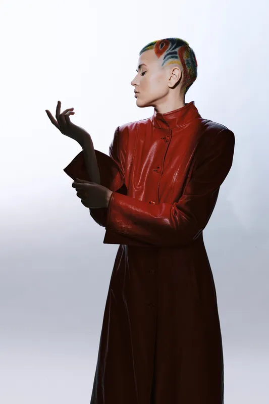 woman with colorful short hair wearing a vibrant red coat, posing sideways against a light background in a modern fashion style, creative and bold look