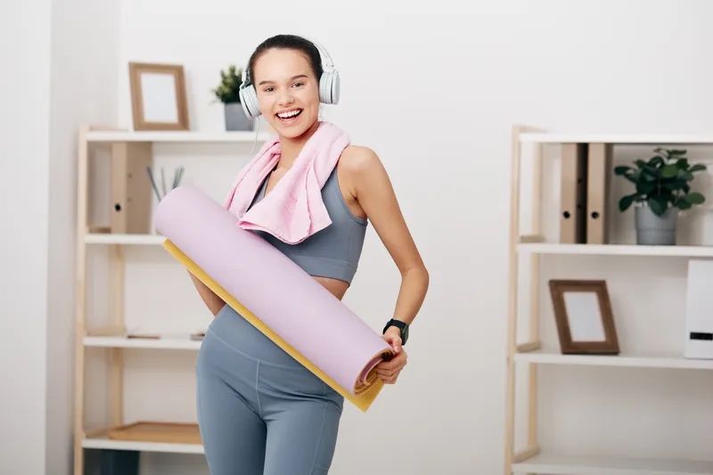 A woman fitness enthusiast in a bright home gym, wearing athletic gear and headphones, holding a yoga mat with a towel; clean room shelves and plants create an inviting space for a full-body workout.