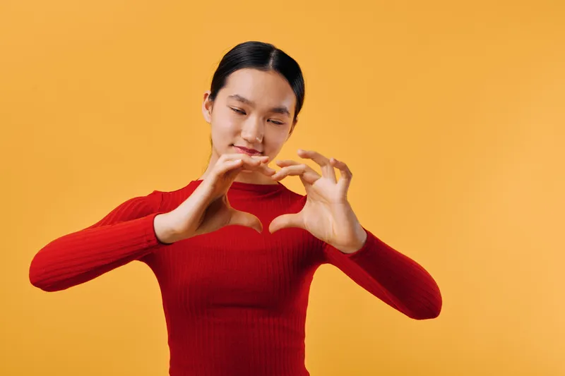 Woman with closed eyes making heart shape with hands in front of orange background for love, affection and emotion. Female in red sweater expressing positivity and warmth.