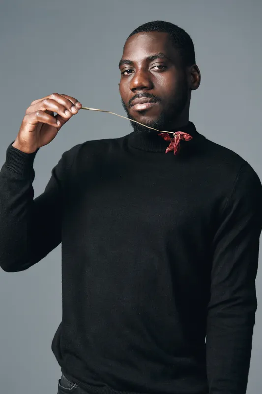 African man in black turtleneck holding fork with red food close to mouth in creative conceptual visual style. Portrait of confident male with dark skin on gray background.