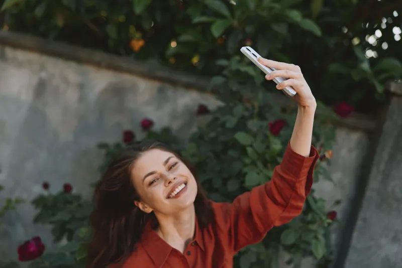 Young woman with long brunette hair wearing a red shirt takes a playful outdoor selfie, smiling brightly as a flowering garden forms a colorful frame