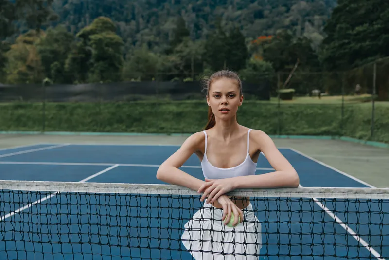 Young athletic woman in sportswear holding a tennis ball, posing confidently on a tennis court surrounded by nature, exuding determination and focus