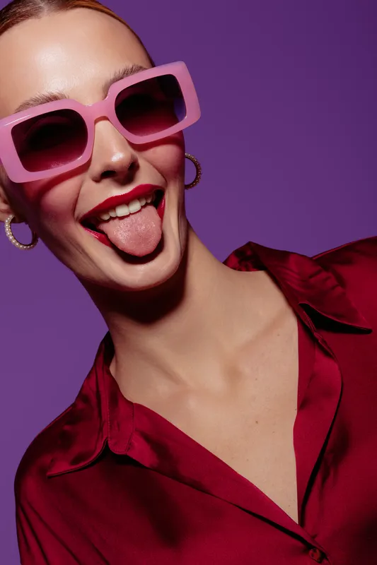 Young woman in pink sunglasses and red shirt sticks out tongue playfully against purple background. Concept of fun, fashion, youth, and carefree attitude in studio portrait.