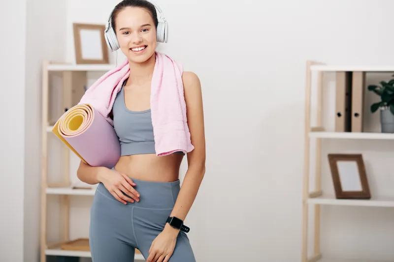 Fitness woman in athletic wear stands with a yoga mat and pink towel in a bright home gym, wearing headphones and ready for a workout session.