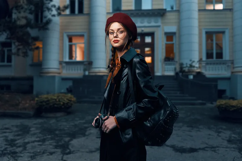 Стильная женщина в красном берете и очках принимает эффектную позу на фоне залитого солнцем исторического здания