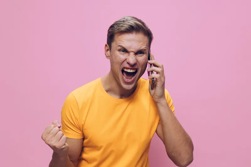 Excited young caucasian man wearing orange t-shirt talking on smartphone clenched fist expression communication digital detox concept bright pink background