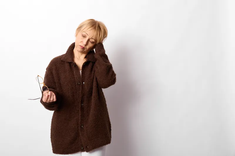 Stressed adult woman in winter coat rubs her head with one hand, holding glasses, portrait isolated on white background, headache and health worry concept