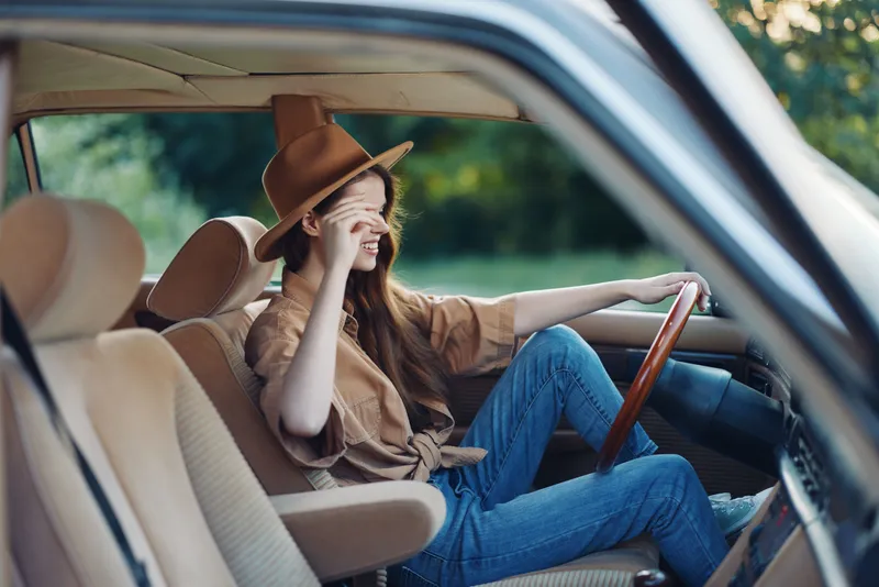 Young woman driving a vintage car, wearing a stylish hat and casual outfit, smiling with joy, in a greenery-filled background.