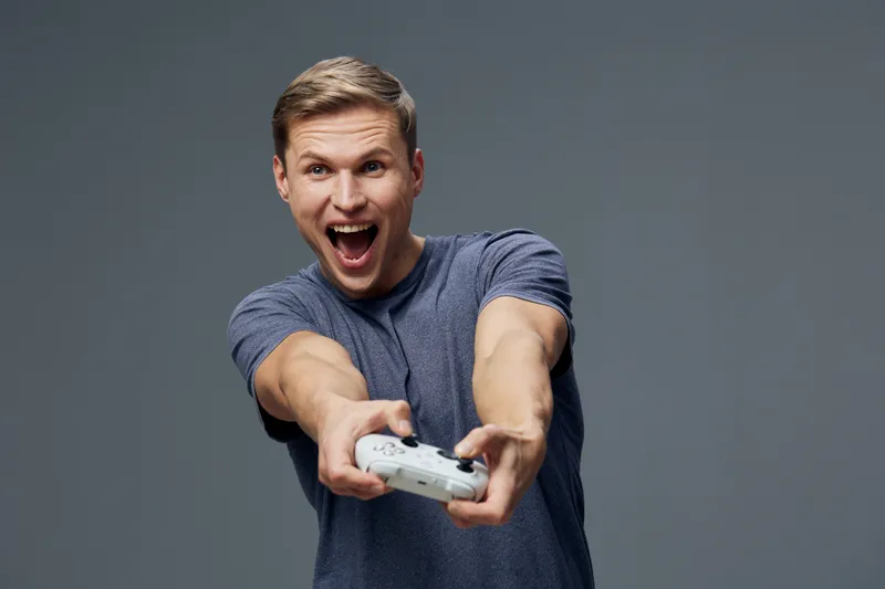 Excited young man with short brown hair wearing casual blue t-shirt enthusiastically playing video game with white controller, expressing joy and surprise, isolated on plain gray background in studio 