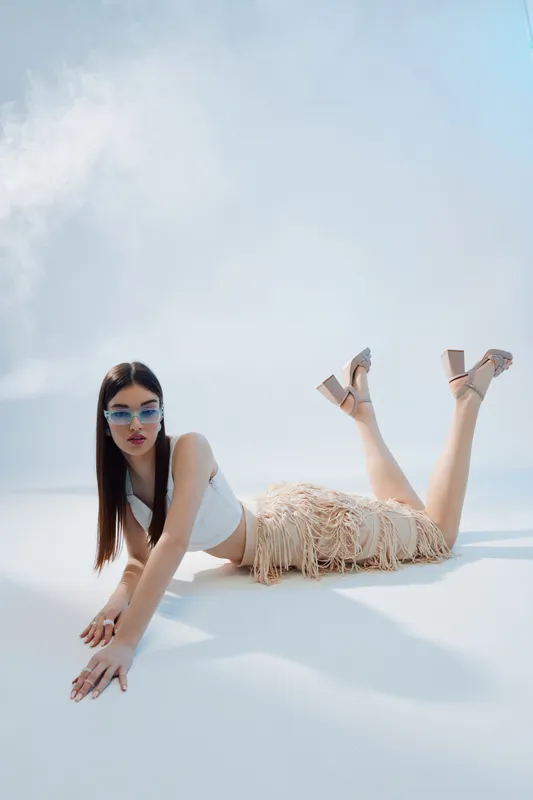 Young woman in stylish sunglasses and fringed skirt poses on the floor in a bright studio. Concept of beauty, fashion, and modern summer style with confident attitude.
