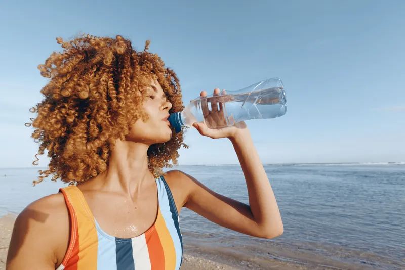 Woman with curly hair drinking water outdoors by the sea, sunlight on face, summer beach setting, relaxed posture, active today, hydration focus, refreshing sip, warm breeze, clear sky