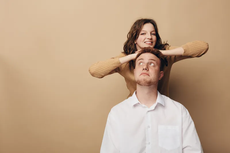 Young man in white shirt looks up thoughtfully while woman in brown sweater smiles behind him with hands on his head. Concept of friendship, trust, and playful interaction.