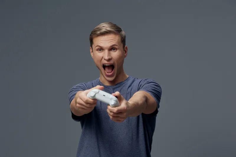 Young enthusiastic man playing video game with controller, expressing excitement and joy, on plain gray background, studio shot, lifestyle concept, casual clothing, male, entertainment.