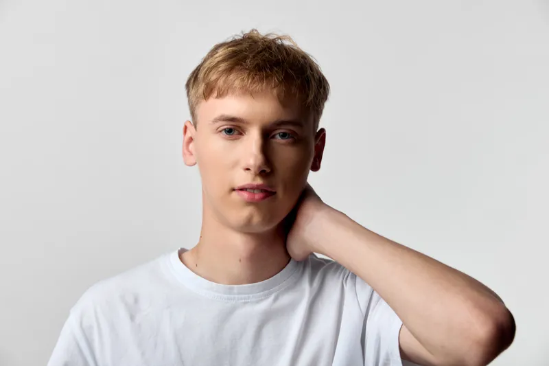 Man with light hair wearing a white t shirt stands against a plain studio background, touching his neck in a casual pose, conveying simplicity, relaxed mood, and contemporary style for lifestyle or