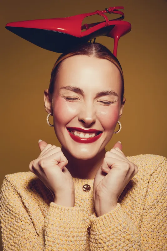 Happy woman with red lipstick and closed eyes smiles joyfully while balancing red high heel shoe on her head. She wears yellow sweater and gold hoop earrings on brown background.