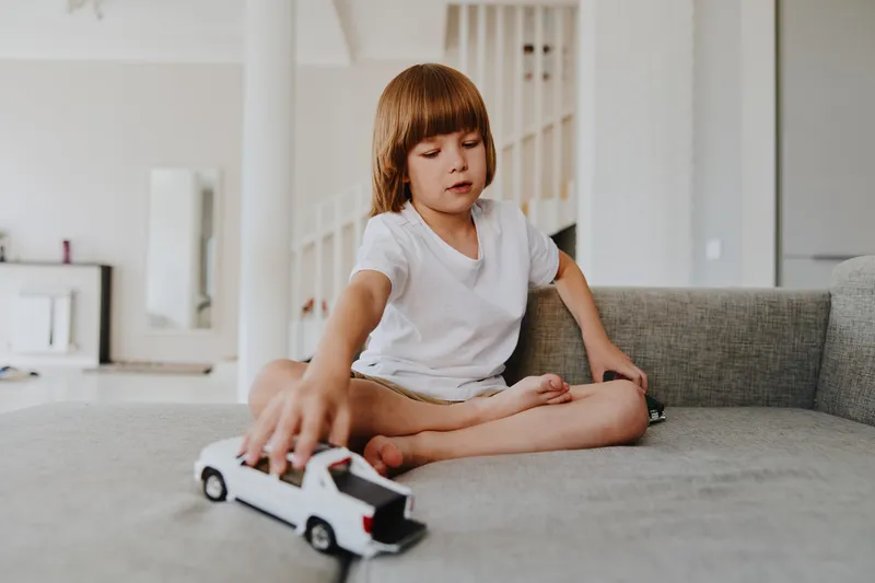 A focused boy playing with a toy car, sitting comfortably on a stylish sofa in a bright and modern living room, with a relaxed atmosphere and playful vibe.