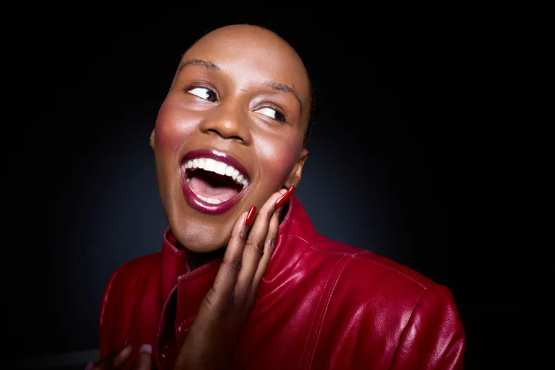 Happy black woman with dark skin wearing red leather jacket smiles widely looking to side. Studio portrait with dark background. Concept of joy, fashion, beauty, and confidence.